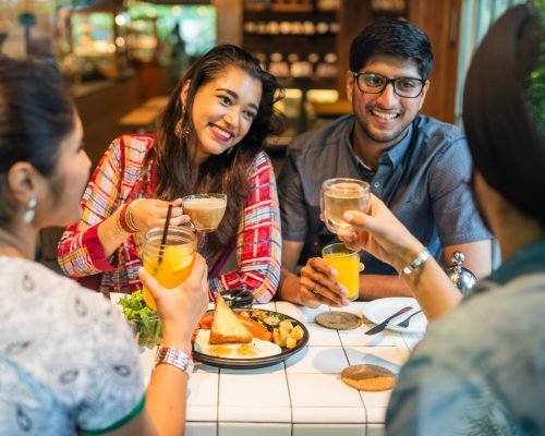 group friends enjoying drinks food smiling chatting they raise glasses toast diverse group relaxed atmosphere cheerful gathering young indian friends eating lunch restaurant scaled